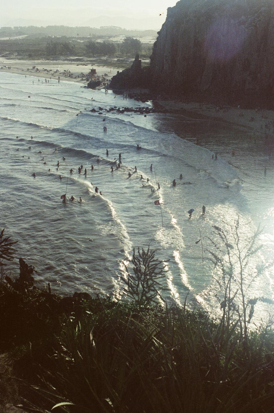 A high-angle view shows a beach with many people in the water and on the sand, next to a large rock formation.