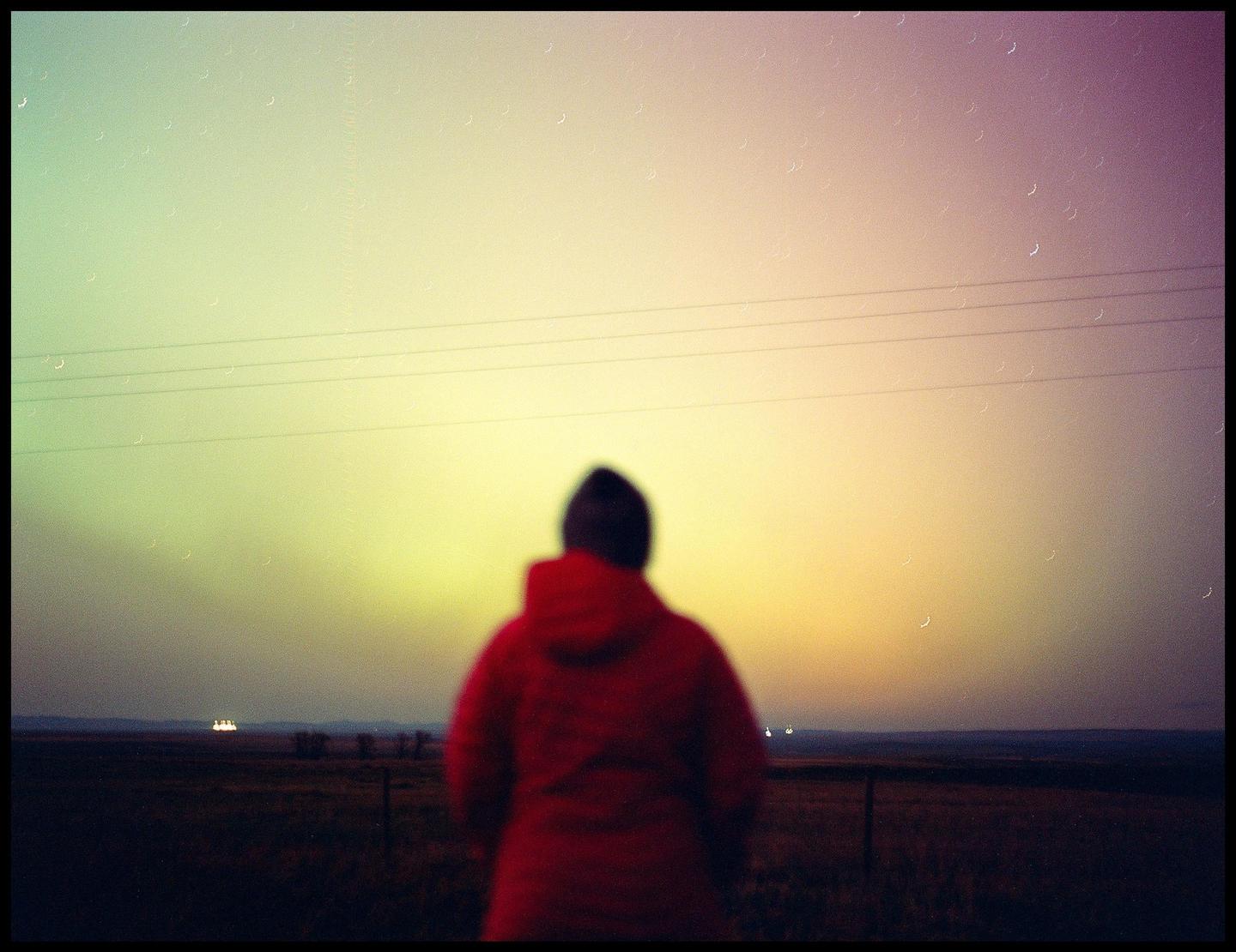 A person in a red jacket stands with their back to the camera, looking out at a colorful sky over a field.