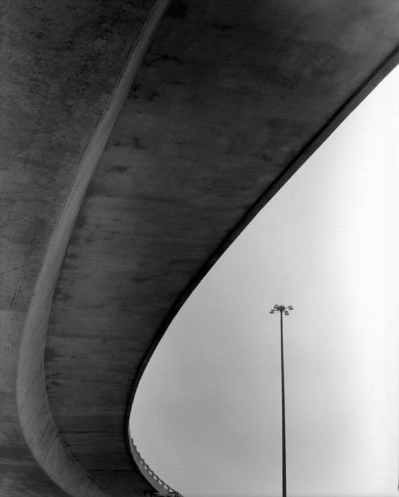A concrete overpass curves over a tall light pole against a bright sky.