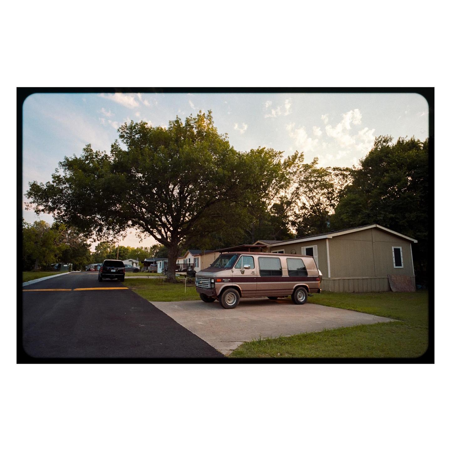 A brown van is parked on a concrete slab next to a house with a large tree in the background.