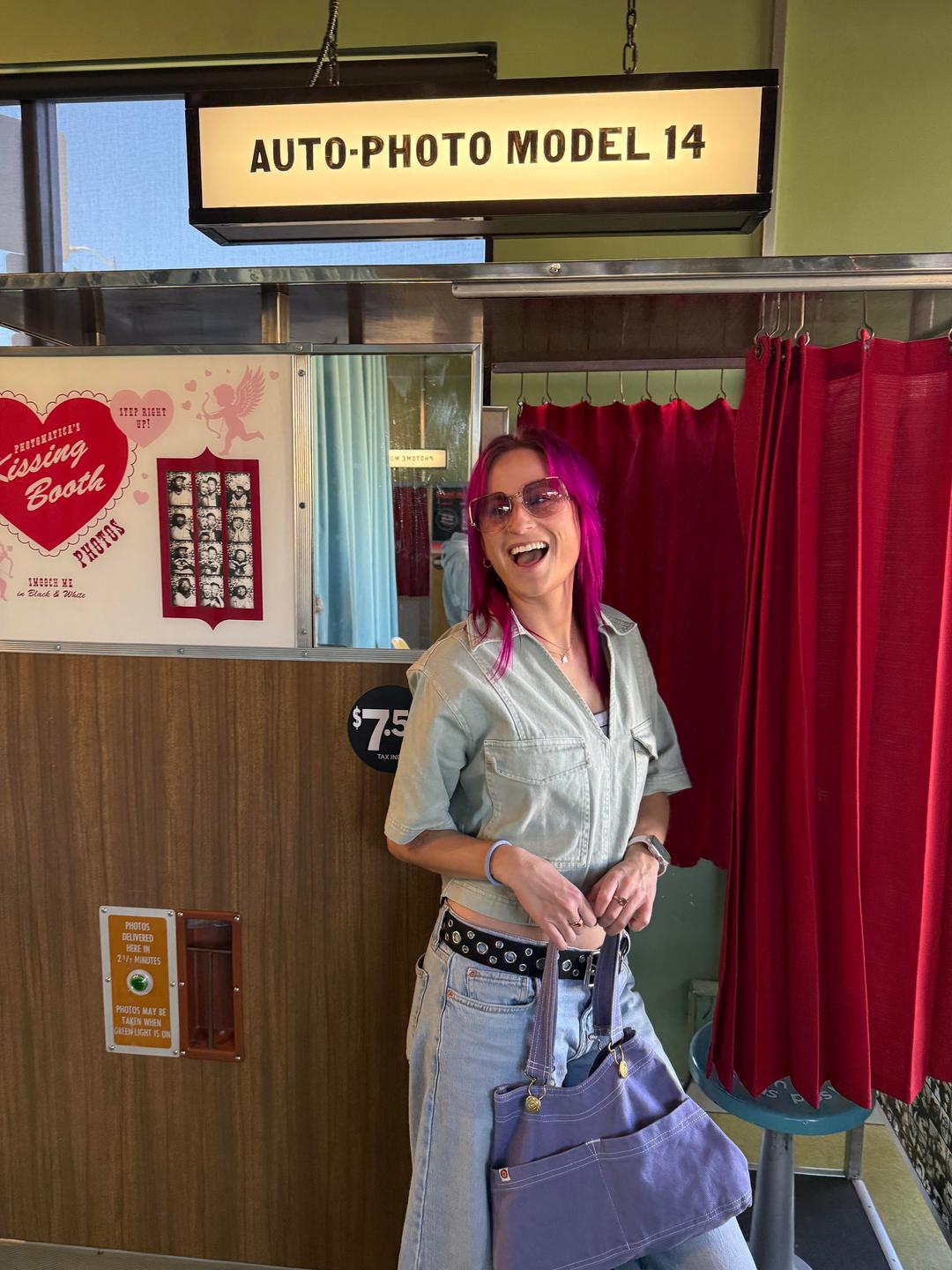 A woman with pink hair and sunglasses stands in front of a vintage photo booth.