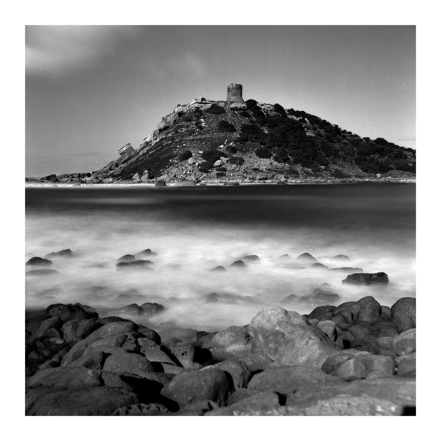 A stone tower stands atop a rocky hill overlooking a body of water with blurred waves and rocks in the foreground.