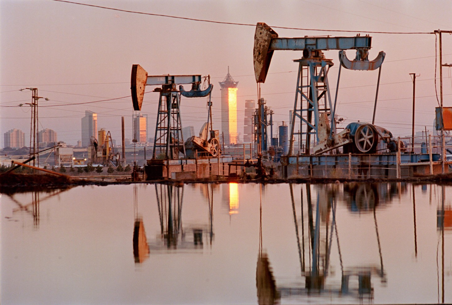 Two oil pumps stand in front of a city skyline with a golden skyscraper reflecting in the water at dusk.