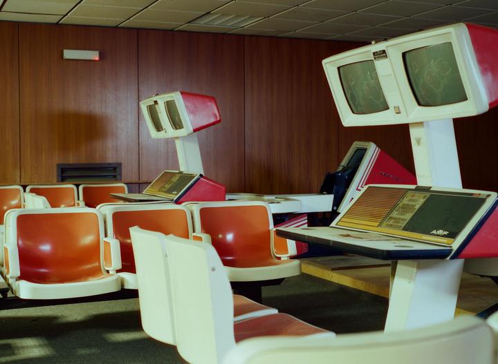 A row of orange and white seats faces bowling alley scoring machines with screens.