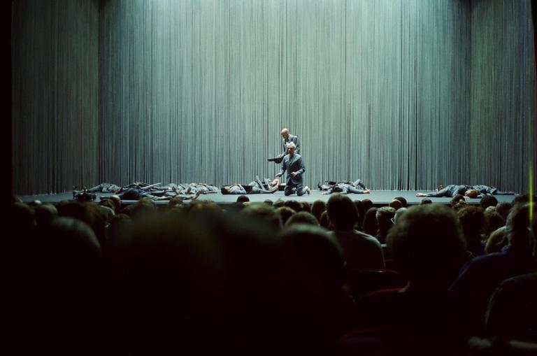 Two men in suits stand on a stage with several other men lying on the floor, viewed from the perspective of an audience member.