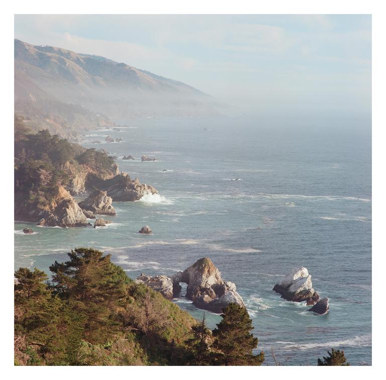 A hazy coastal landscape shows a rugged shoreline with waves crashing against rocks and mountains in the distance.