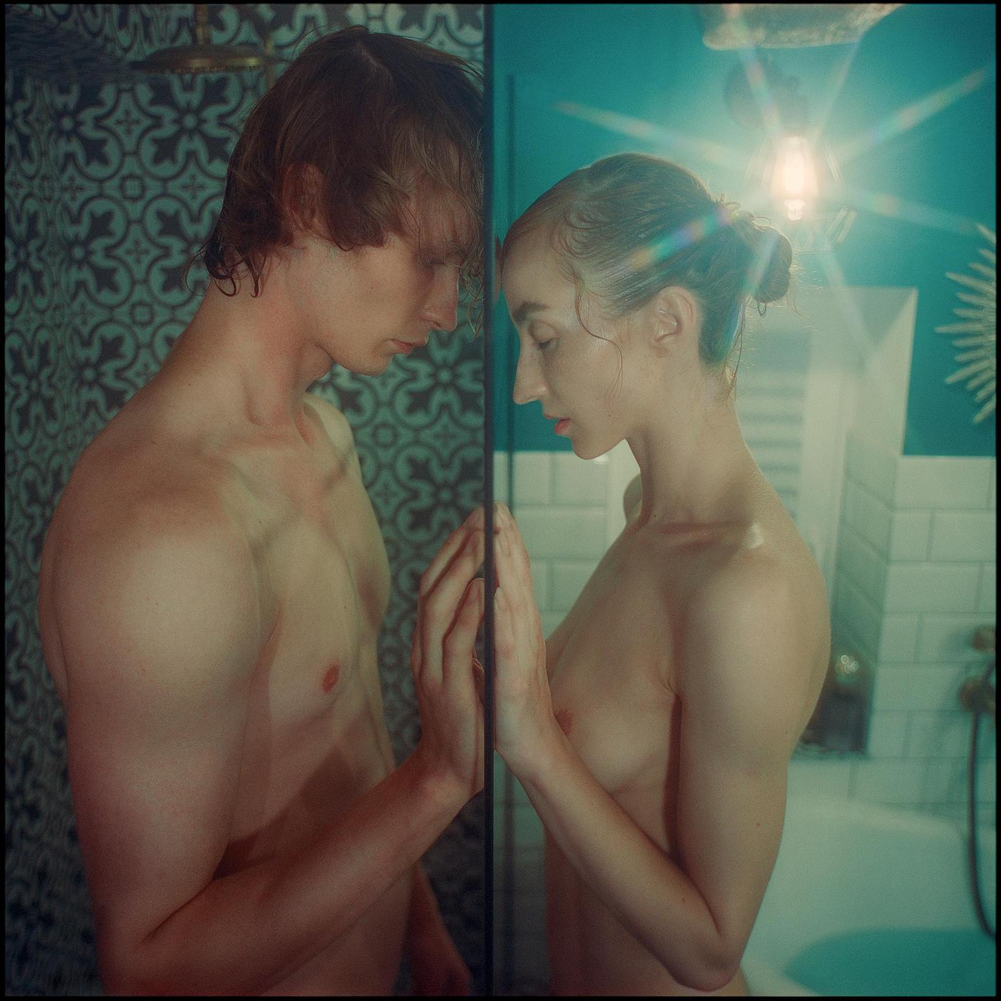 A man and a woman stand on opposite sides of a glass shower door, touching hands.