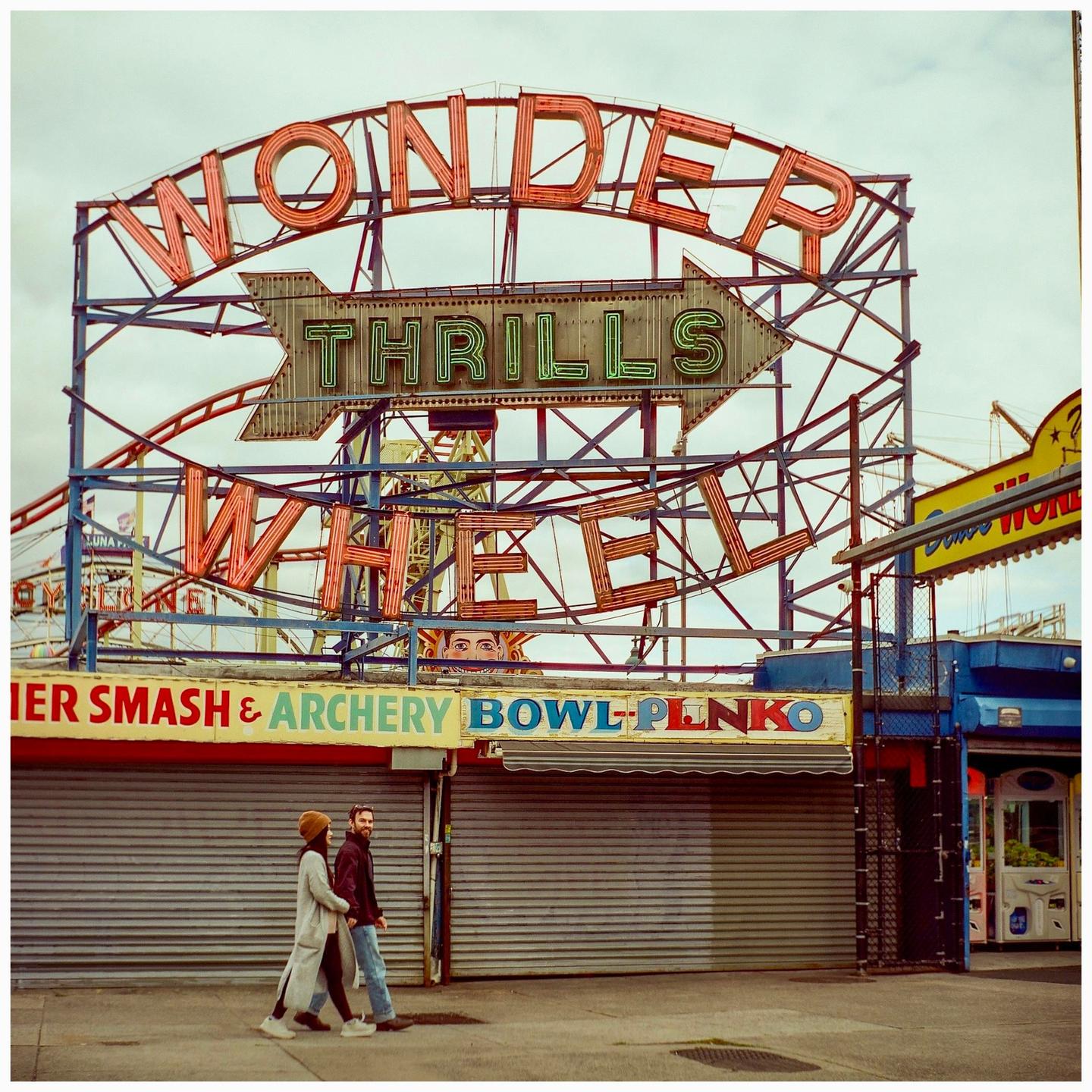 A couple walks past a large neon sign for "Wonder Wheel Thrills" at an amusement park.