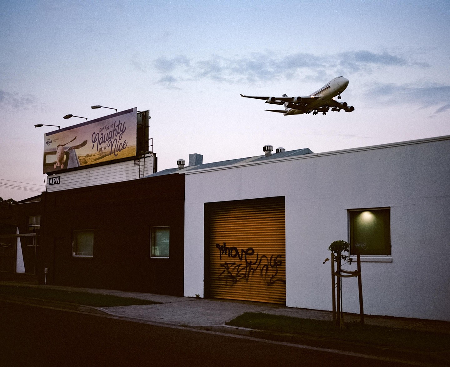 A large airplane flies low over buildings with a billboard and a garage door with graffiti.
