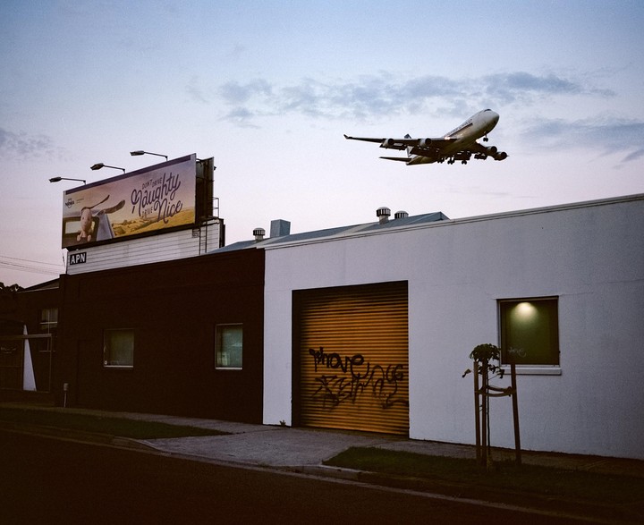 A large airplane flies low over buildings with a billboard and a garage door with graffiti.