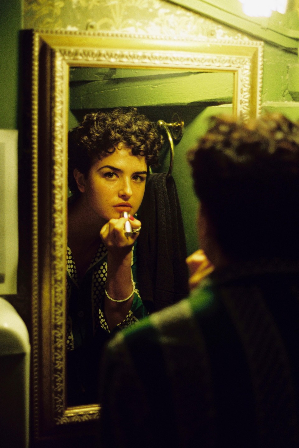 A woman with curly hair applies lipstick while looking into a mirror.