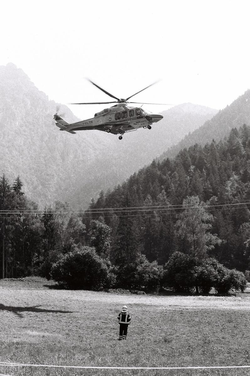 A helicopter takes off over a field where a person in uniform stands, with mountains and trees in the background.