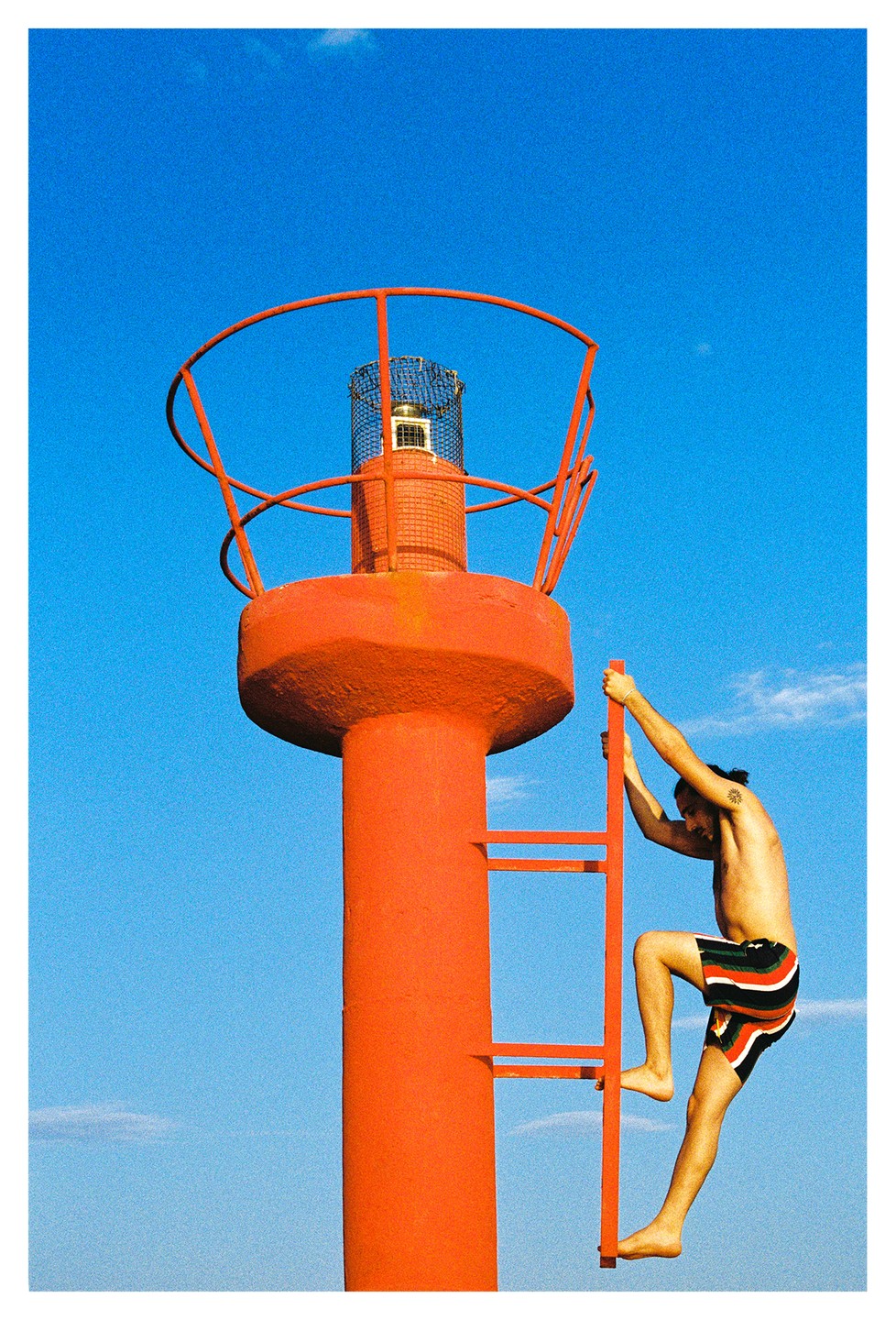 A man climbs a red ladder on a red lighthouse against a clear blue sky.