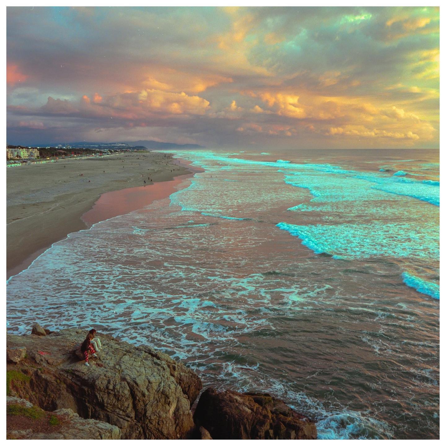 A person sits on a rocky cliff overlooking a beach with waves crashing on the shore under a dramatic sky.