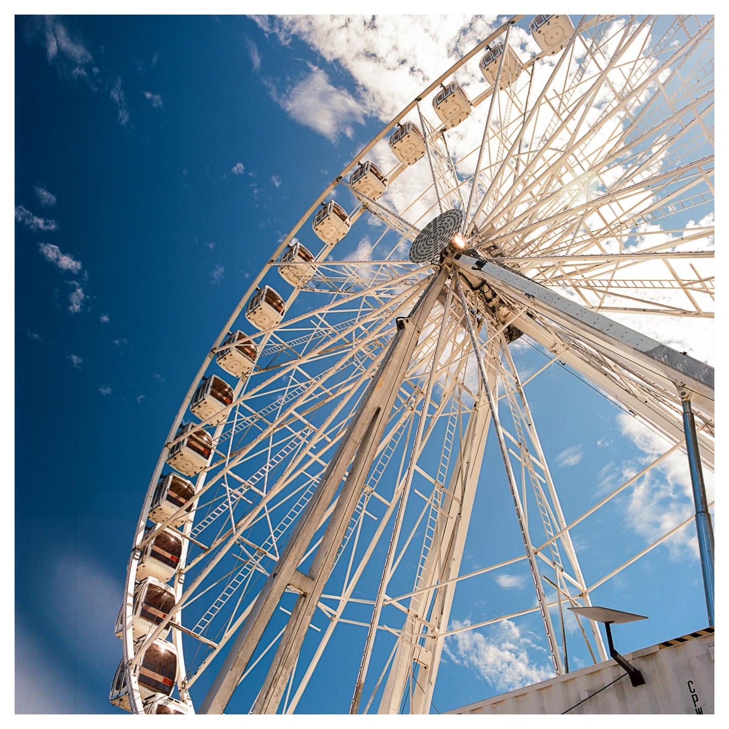 A low-angle view shows a large white ferris wheel against a bright blue sky with scattered clouds.