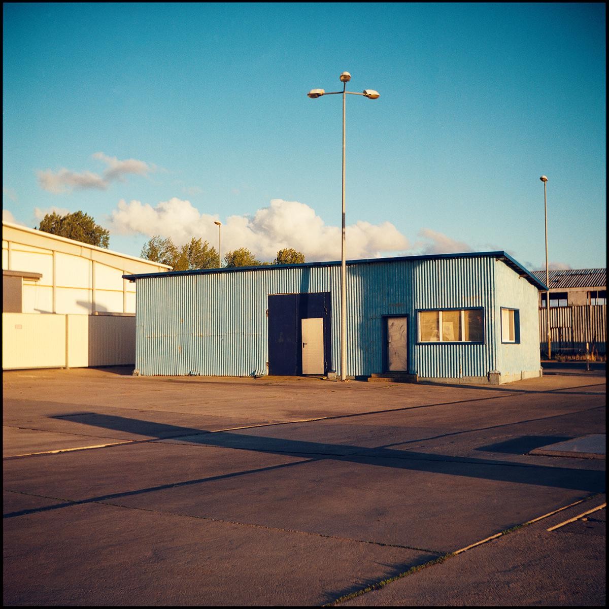 A blue corrugated metal building with a tall street light in front of it stands on an asphalt lot under a blue sky with white clouds.