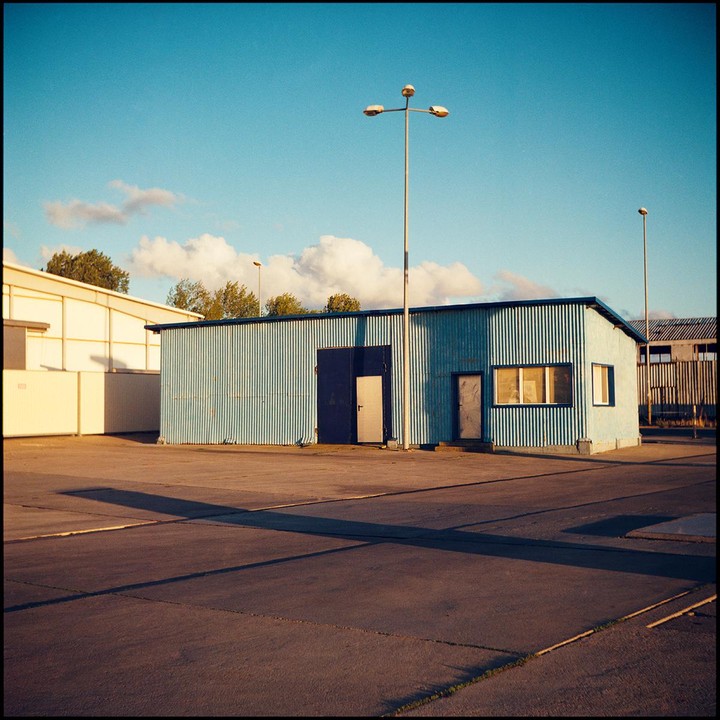 A blue corrugated metal building with a tall street light in front of it stands on an asphalt lot under a blue sky with white clouds.