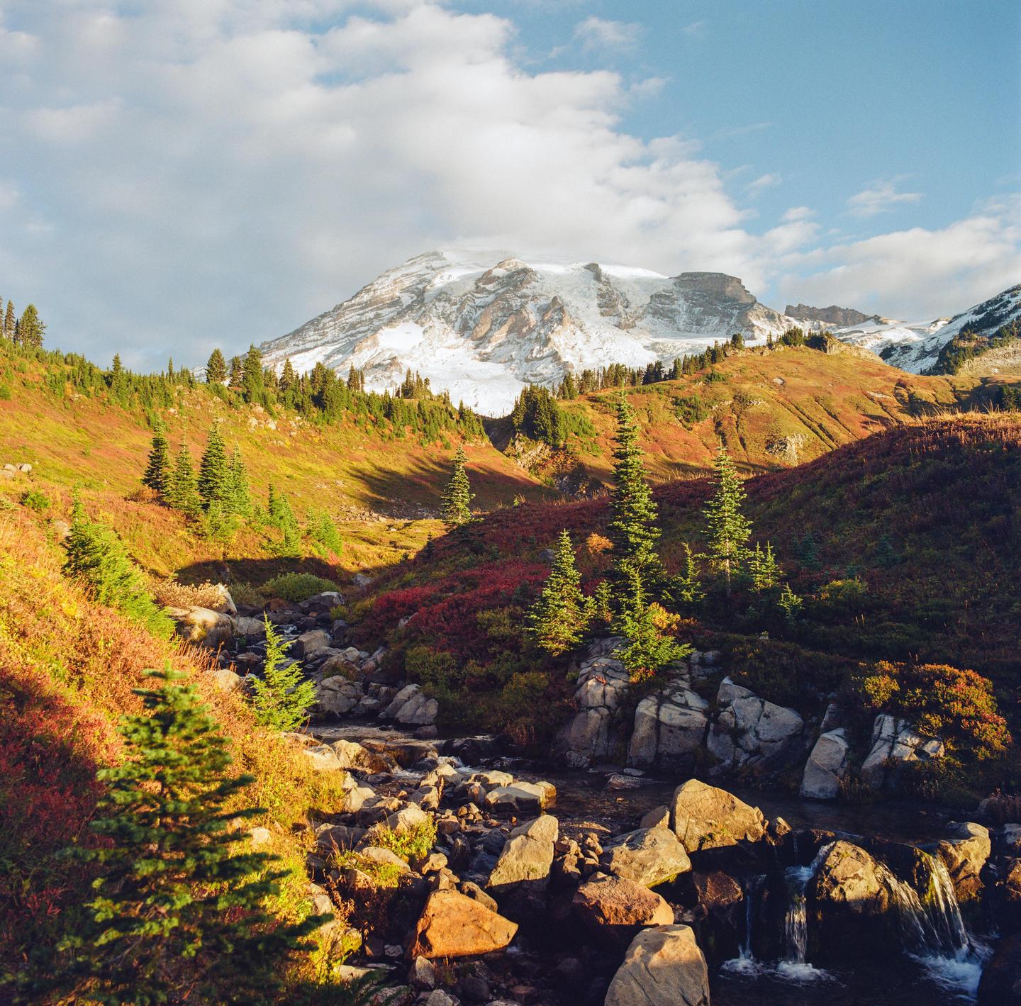 A snow-capped mountain rises above a valley with a rocky stream and autumn foliage.