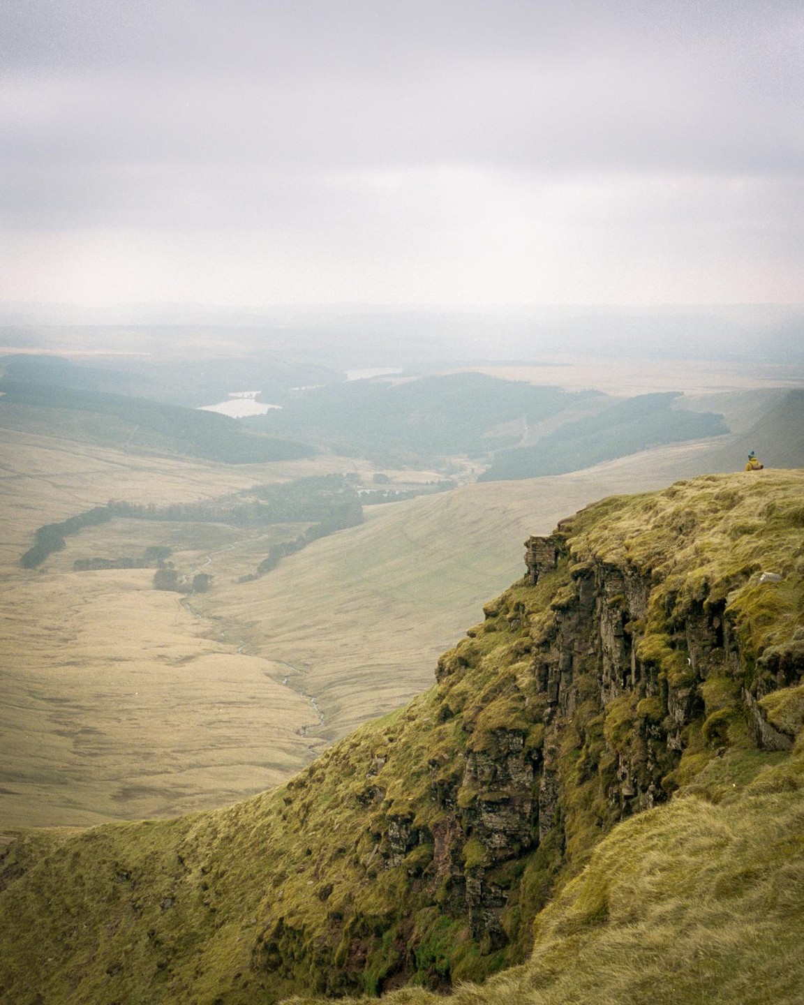 A person sits on a grassy cliff overlooking a vast valley with a lake in the distance under a hazy sky.
