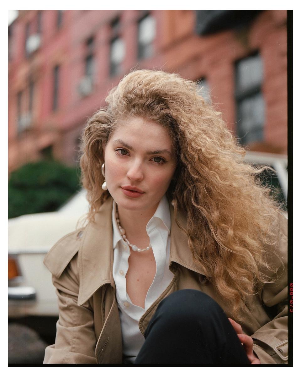 A woman with long curly hair and a trench coat looks at the viewer.