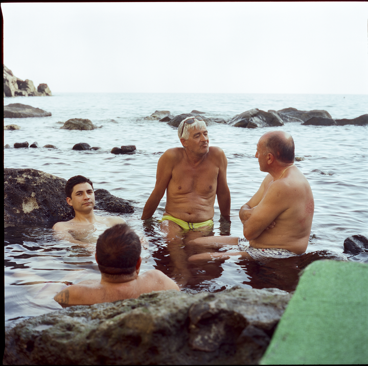 Four men are sitting in the water among rocks at a thermal bath.