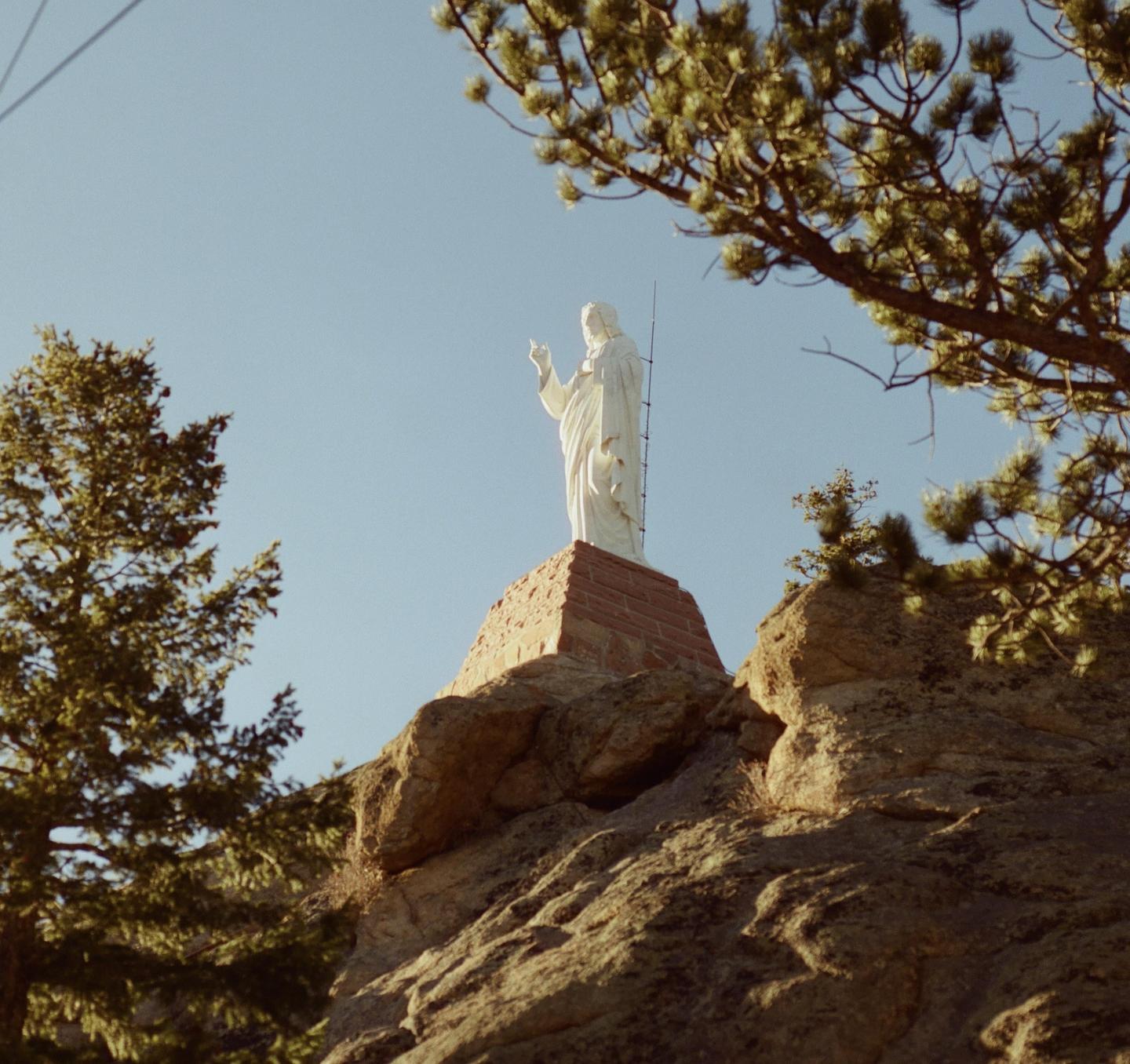 A white statue of Jesus stands on a brick pedestal atop a rocky hill, framed by pine trees against a clear sky.