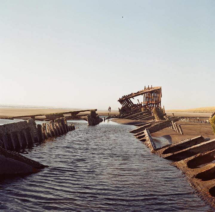 A shipwreck lies on a sandy beach with a small stream of water flowing towards the ocean.