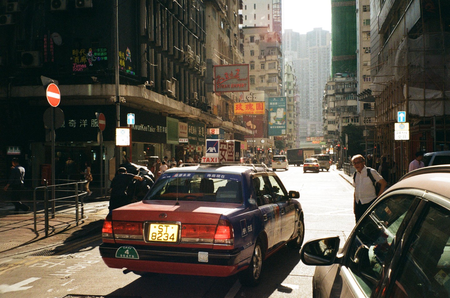 A red and blue taxi is parked on a busy street in Hong Kong with tall buildings and many signs in the background.