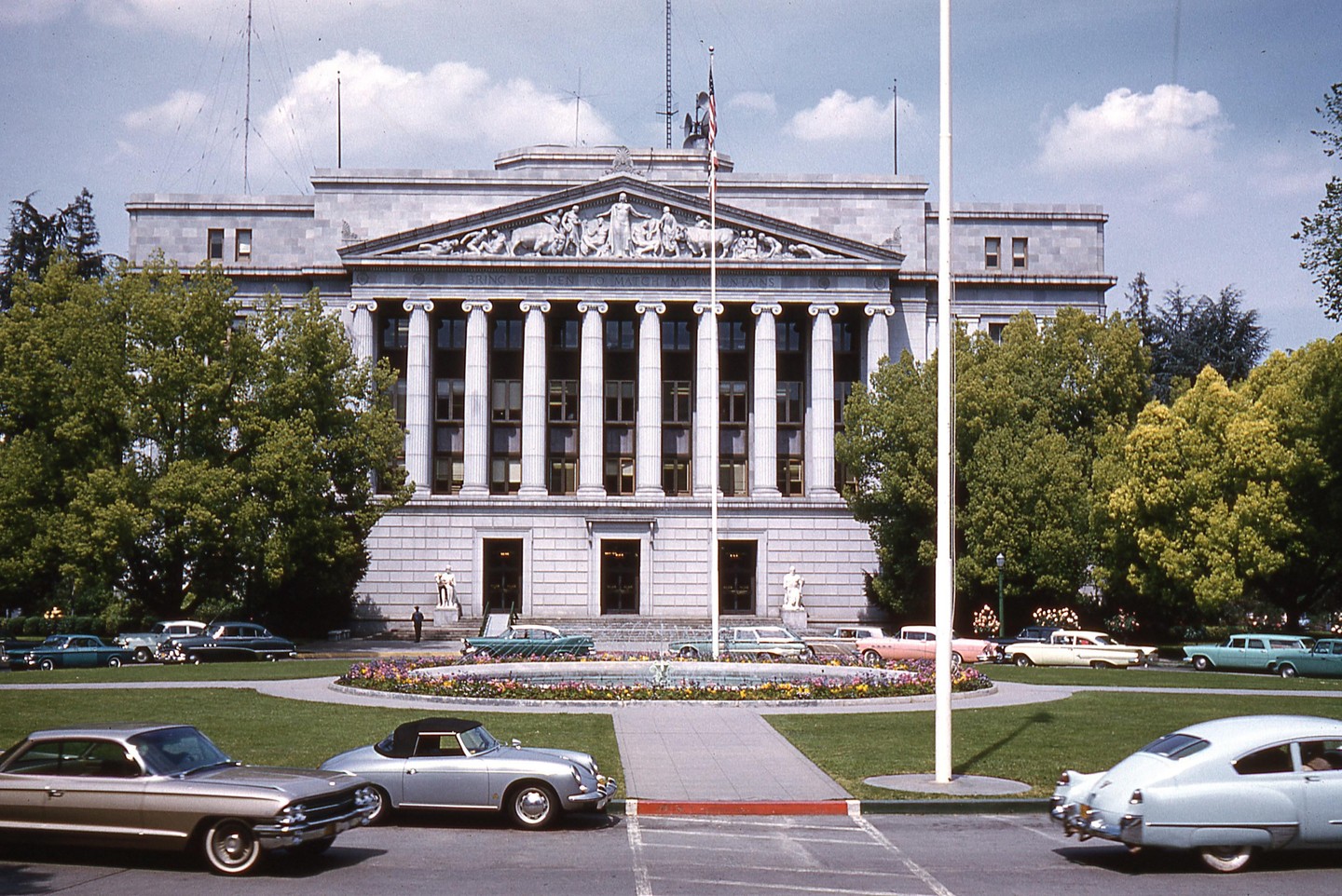 A large stone building with columns and a decorative facade stands behind a circular flower bed and several vintage cars.