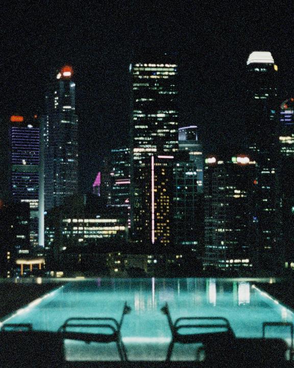 A city skyline at night is reflected in a swimming pool with two chairs in the foreground.