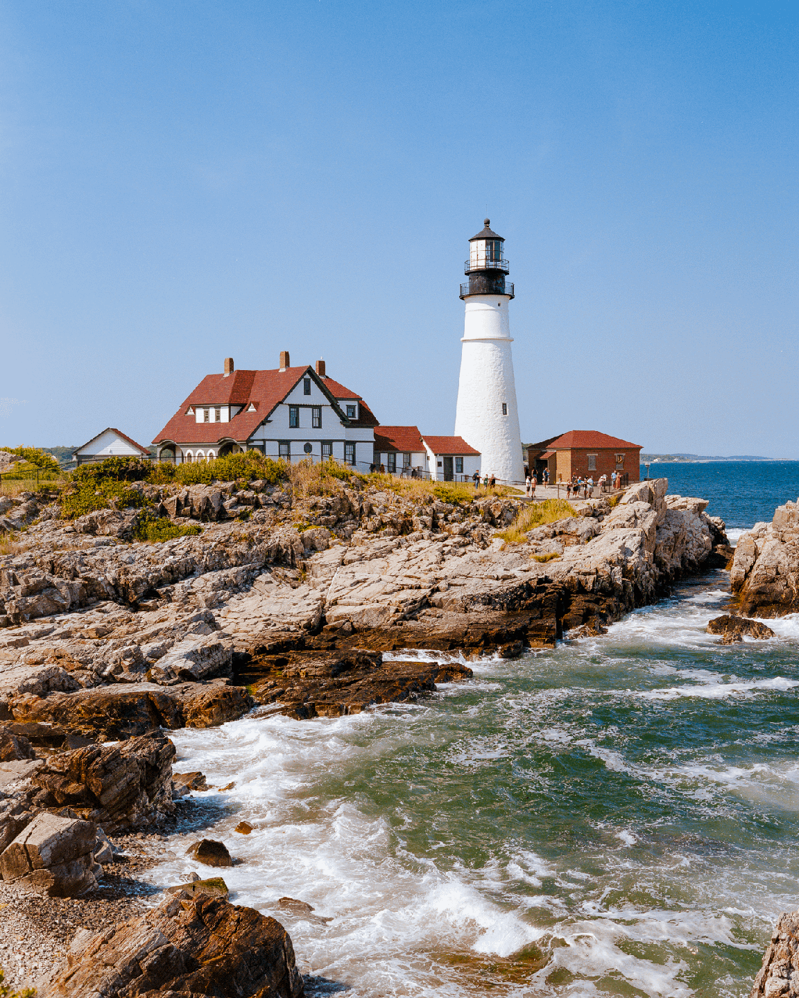 A white lighthouse stands on a rocky coast next to a building with a red roof, with waves crashing against the shore.