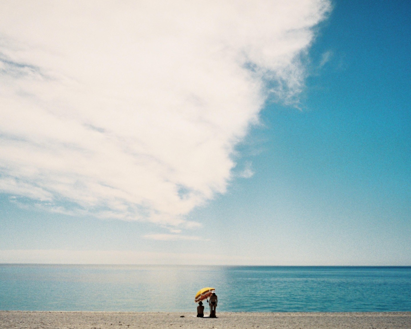 Two people stand under a yellow umbrella on a beach next to the ocean under a partly cloudy sky.