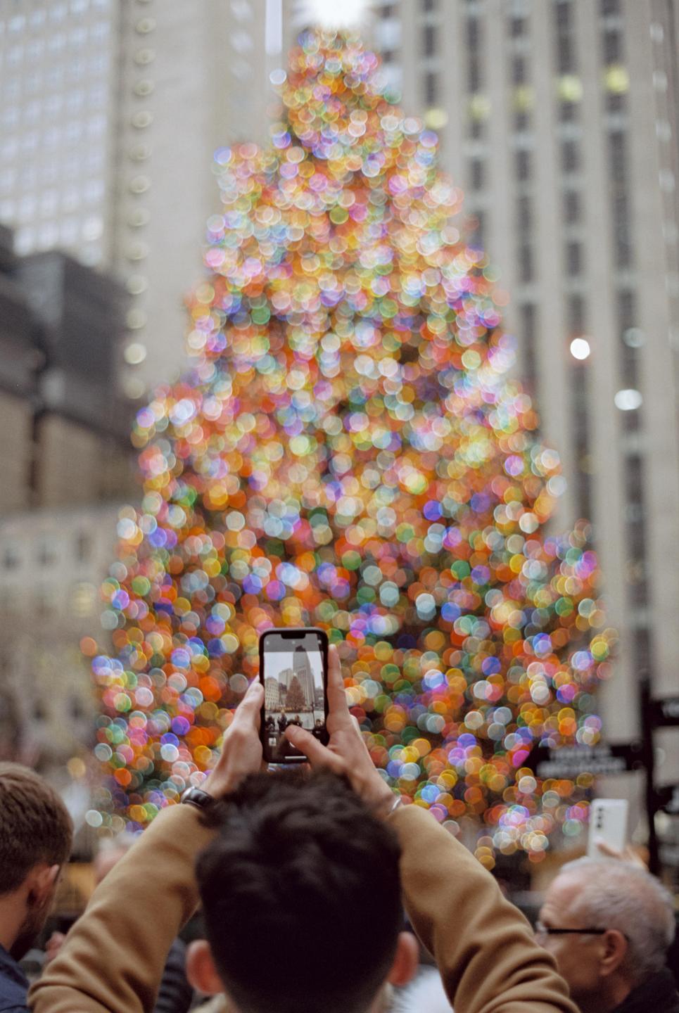 A person holds a phone to photograph a large, out-of-focus Christmas tree made of colorful lights.