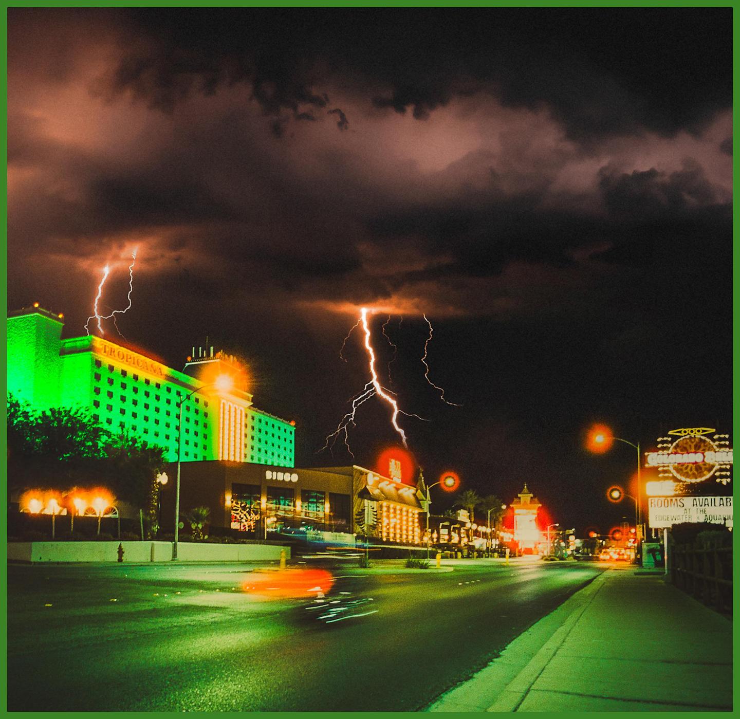Lightning strikes over a street with illuminated buildings and blurred car lights at night.
