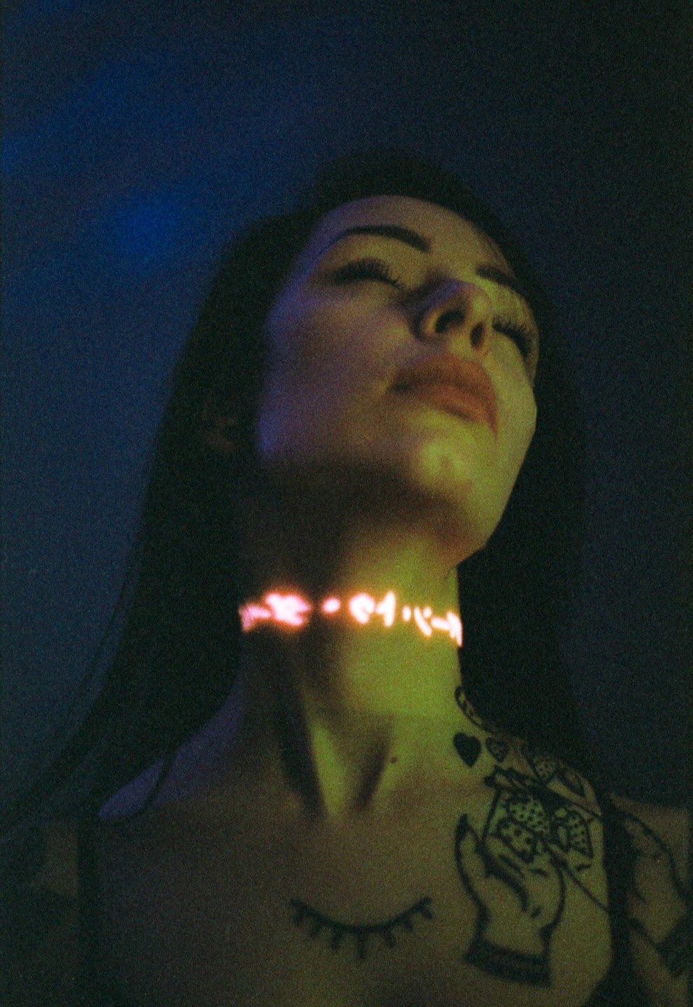 A woman with closed eyes has glowing Japanese characters projected onto her neck and tattoos on her chest and shoulder.