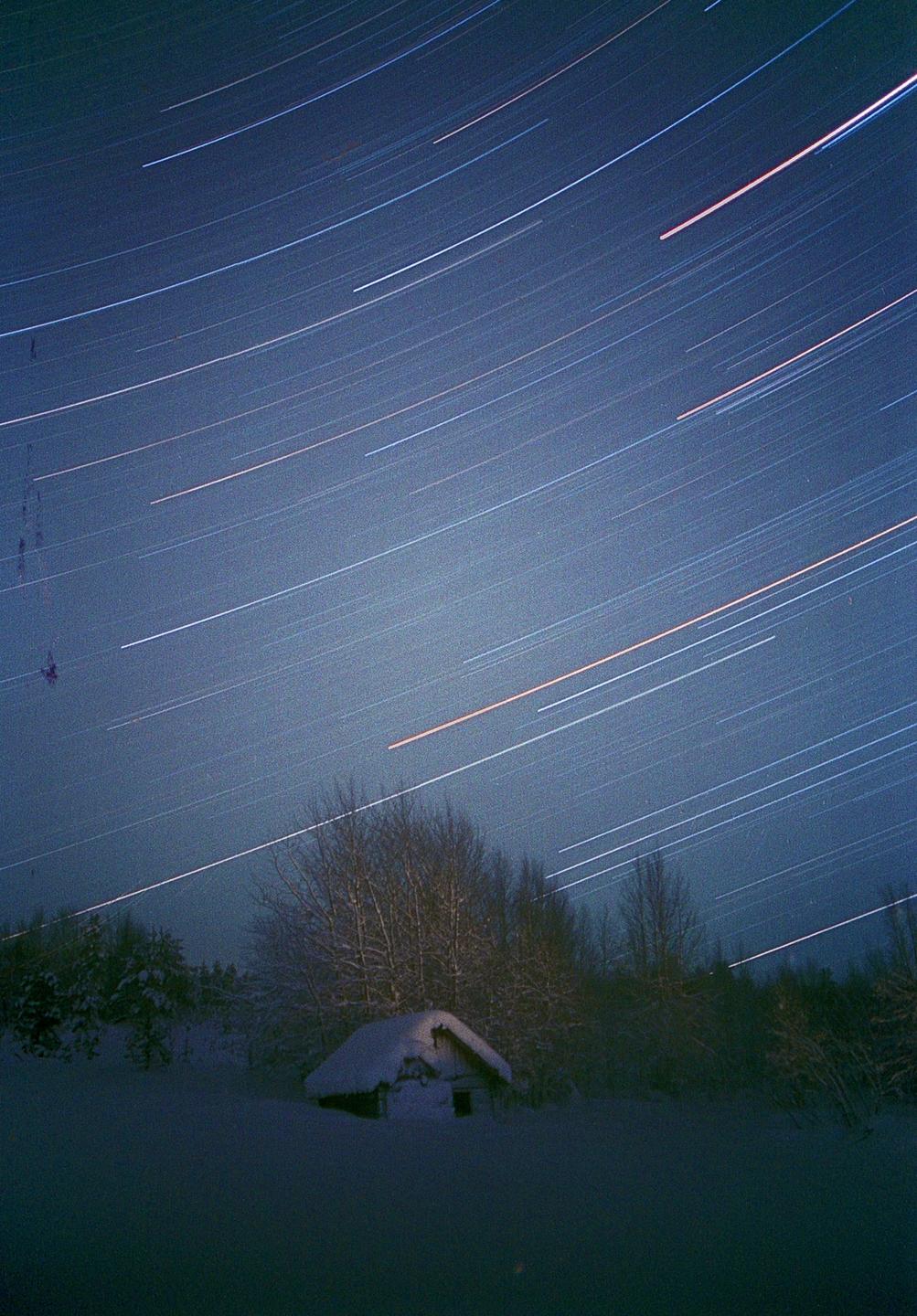 A small snow-covered cabin sits in a snowy landscape under a night sky filled with star trails.