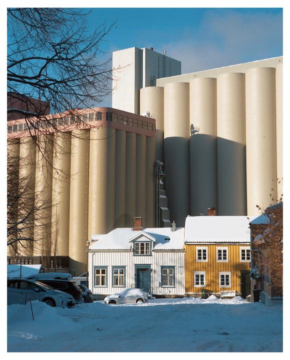 Two snow-covered houses stand in front of large industrial silos under a clear blue sky.