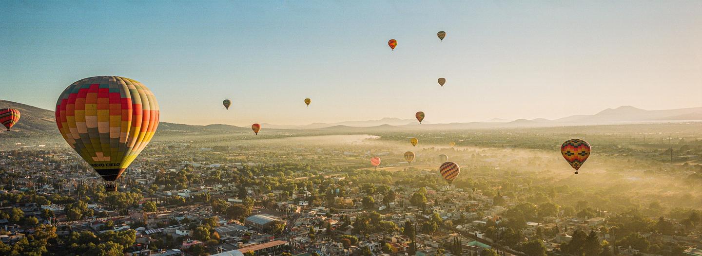 Many hot air balloons float above a hazy town with mountains in the distance.