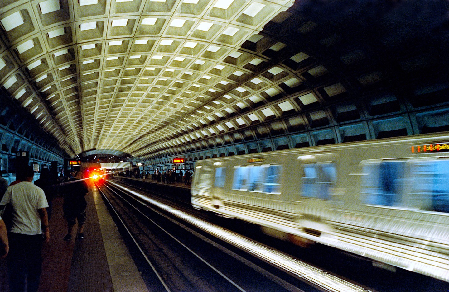 A subway train speeds through a station with a distinctive waffle-patterned ceiling.