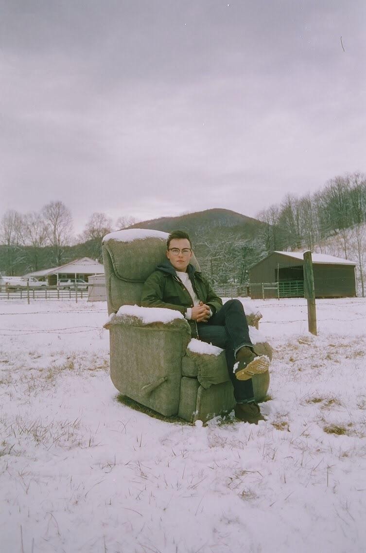 A man sits in a snow-covered armchair in a snowy field with a barn and hills in the background.