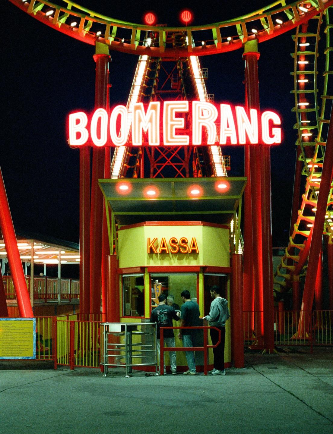 Three men stand in front of a ticket booth at an amusement park ride called Boomerang at night.