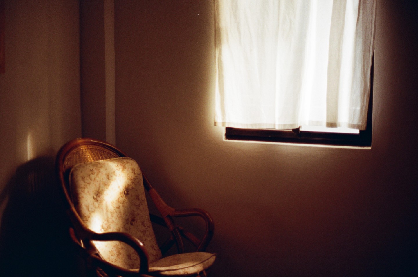 Sunlight streams through a window with a white curtain, illuminating a patterned cushion on a wooden chair in a dimly lit room.