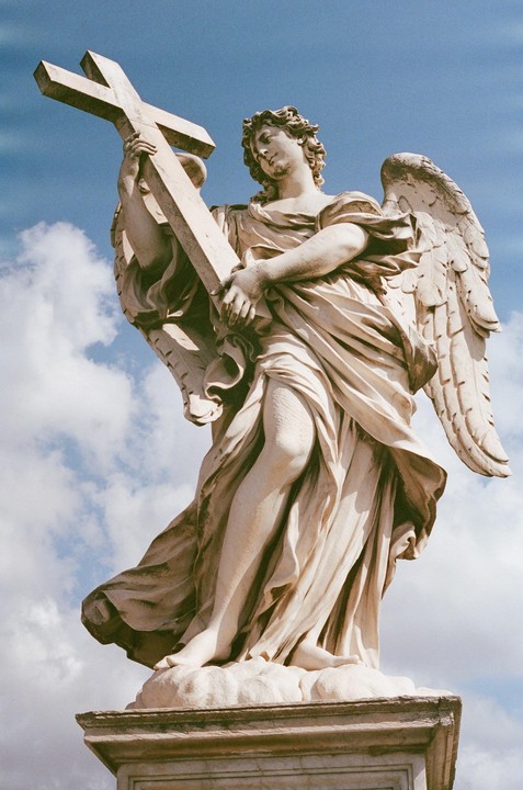 A low-angle shot shows a stone angel statue holding a cross against a blue sky with clouds.