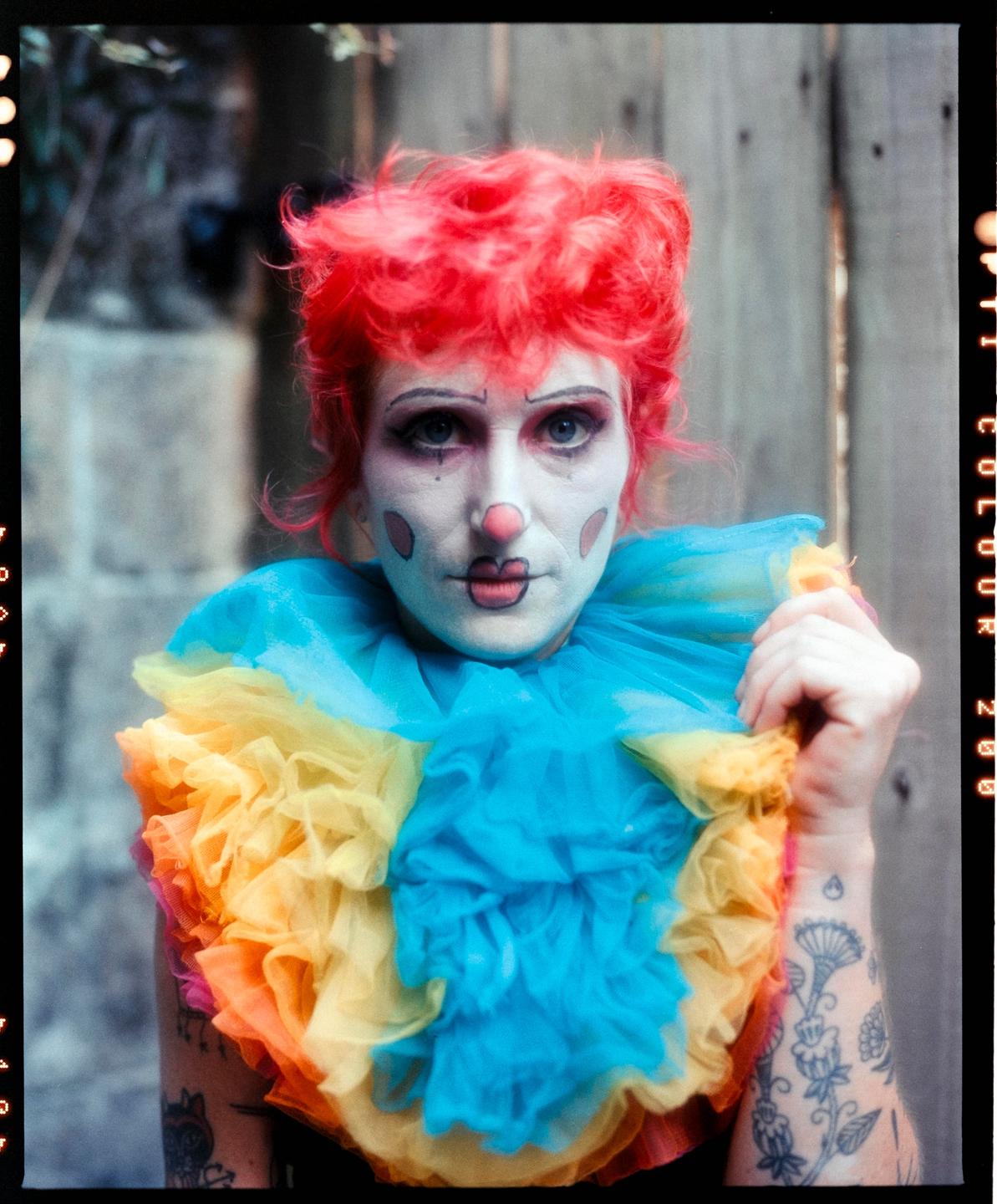 A person with red hair and clown makeup wears a colorful ruffled collar.