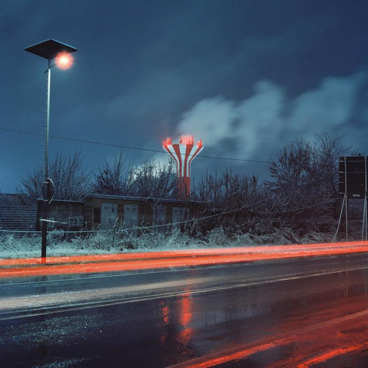 Red light trails from a vehicle pass on a wet road next to a striped water tower emitting smoke at night.