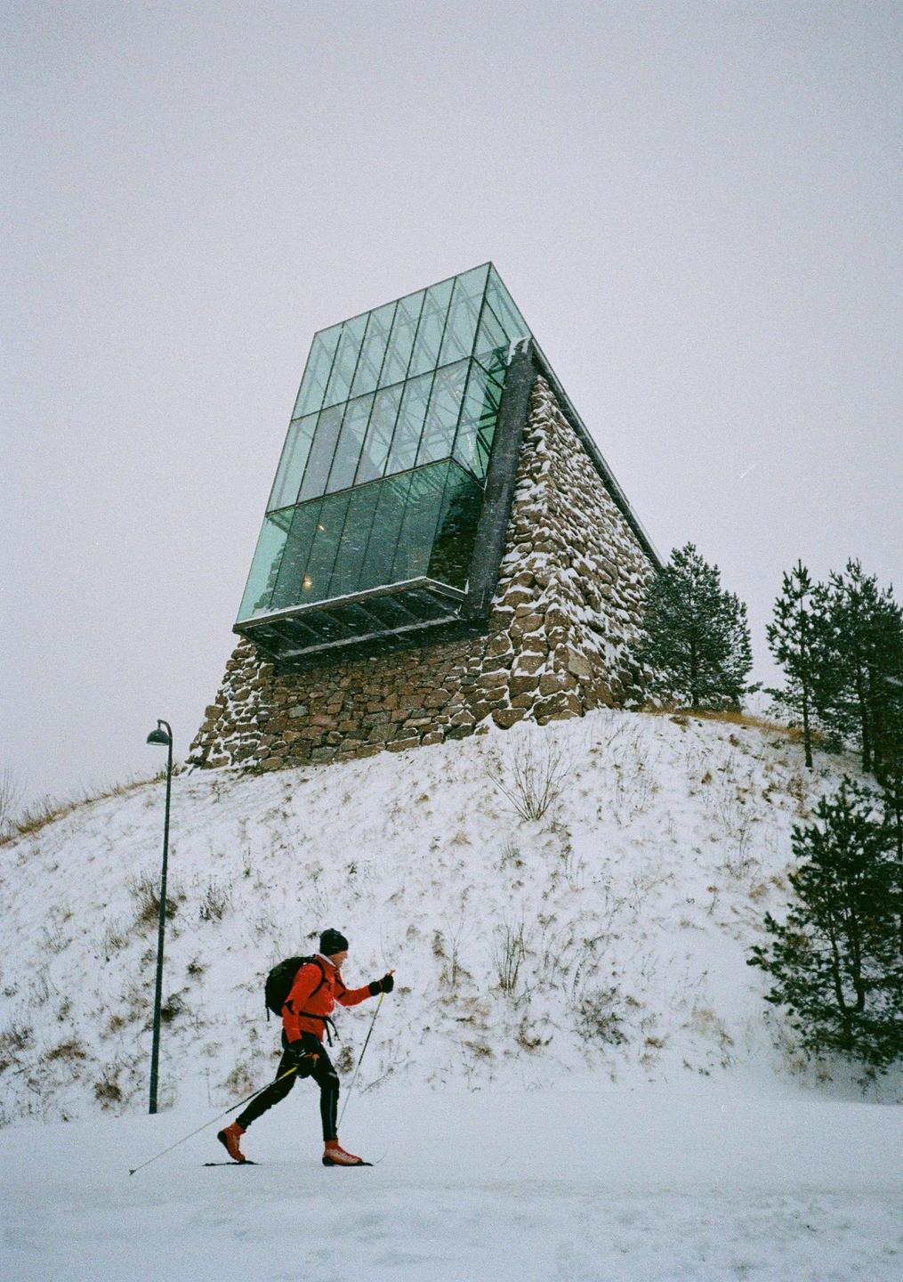 A man cross-country skis in front of a modern building with a glass and stone facade on a snowy hill.