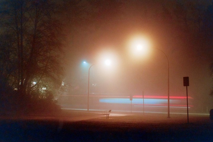 A foggy night scene shows a park bench next to a road with light trails from passing vehicles and streetlights illuminating the mist.