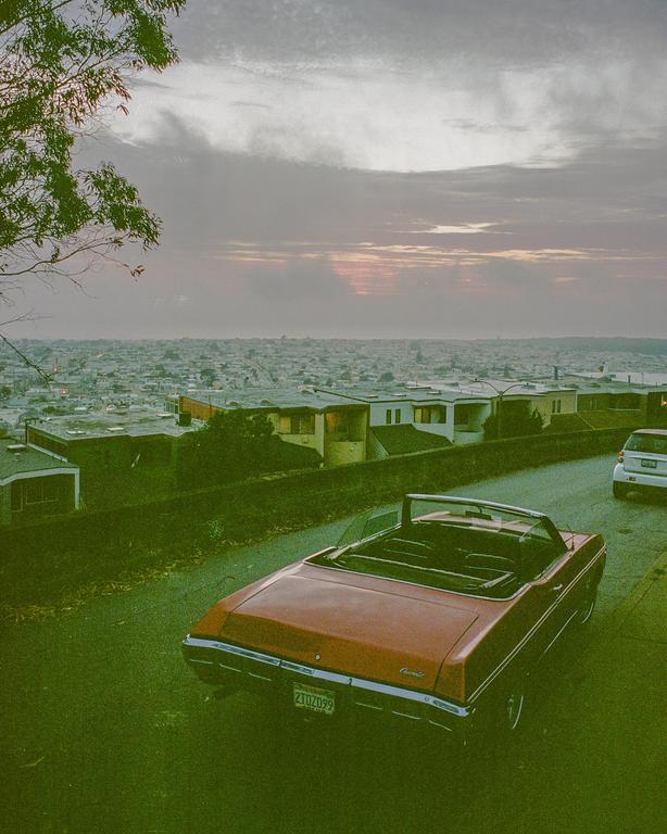 A red convertible is parked on a street overlooking a city at dusk.
