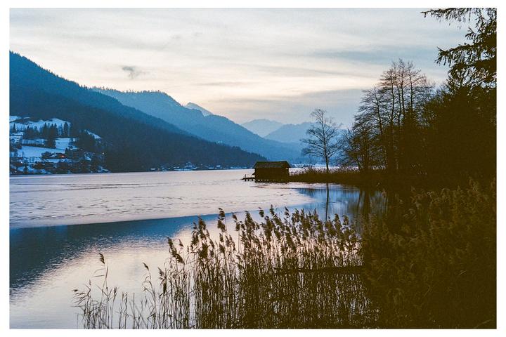 A small wooden cabin sits on the edge of a partially frozen lake, surrounded by mountains and reeds.
