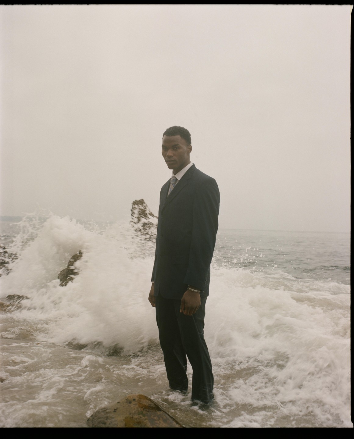 A man in a suit stands in the ocean as waves crash around him on a hazy day.